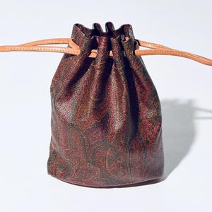 May include: A drawstring pouch with a paisley pattern in shades of burgundy, green, and gold. The bag has a leather drawstring closure and a leather strap. The pouch is approximately 6 inches tall and 5 inches wide.