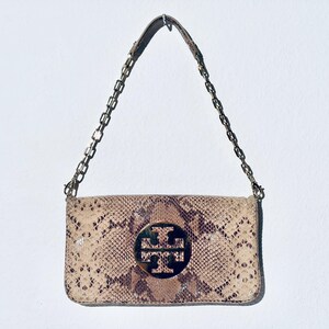 Può includere: Una borsa rettangolare con un motivo a pelle di serpente nei toni del beige e del marrone. Presenta una tracolla a catena color oro e un emblema circolare con un logo al centro. La borsa è sospesa su uno sfondo bianco.