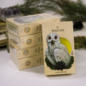 以下が含まれることがあります： 一番上の箱に「The Wooden Tarot」と書かれた箱の積み重ね。箱には、黒い目をした白いフクロウと黒いバラのイラストが描かれている。「70 Card Tarot Deck」という文字が箱の下部に印刷されている。