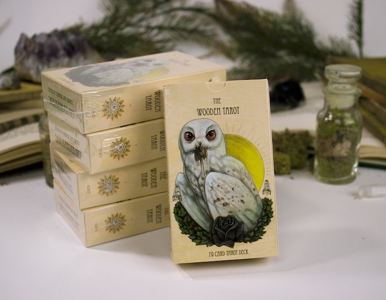 以下が含まれることがあります： 一番上の箱に「The Wooden Tarot」と書かれた箱の積み重ね。箱には、黒い目をした白いフクロウと黒いバラのイラストが描かれている。「70 Card Tarot Deck」という文字が箱の下部に印刷されている。