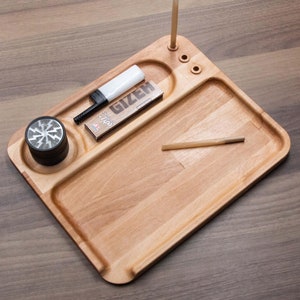 Pode incluir: Uma bandeja de rolar de madeira com um moedor, um pacote de papéis de enrolar, um isqueiro e dois palitos de madeira. A bandeja tem uma borda elevada e um pequeno compartimento para guardar objetos.