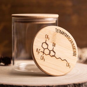 Può includere: Un barattolo di vetro trasparente con coperchio in legno. Il coperchio è inciso con la struttura chimica del tetraidrocannabinolo e la parola "TETRAHYDROCANNABINOL". Il barattolo è su una superficie di legno.
