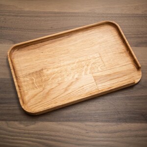 Könnte beinhalten: Ein rechteckiges Holztablett mit abgerundeten Ecken. Das Tablett besteht aus hellem Holz mit sichtbaren Maserungsmustern. Die Kanten sind leicht erhöht und bilden einen Rand um den Umfang. Das Tablett ist leer und scheint neu zu sein.