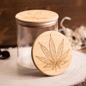 Può includere: Un barattolo di vetro trasparente con un coperchio in bambù con un design a foglia di cannabis inciso. Il coperchio è di lato, rivelando l'interno del barattolo. Il barattolo è su una superficie di legno.