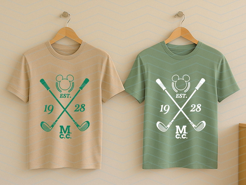 May include: Two short-sleeved t-shirts, one tan and one green, hanging on wooden hangers. Each shirt features a golf-themed design with crossed golf clubs, a Mickey Mouse head, the text "EST. 1928," and "M.C.C."