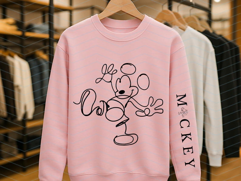 Puede incluir: Una sudadera de cuello redondo en color rosa claro con un dibujo de l&iacute;nea continua negra de Mickey Mouse en la parte frontal. La manga izquierda muestra el texto "MICKEY" impreso verticalmente en una fuente serif negra, con un peque&ntilde;o icono gr&aacute;fico sobre la M.