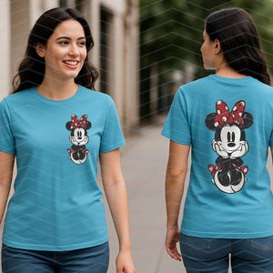 May include: A light blue t-shirt featuring a Minnie Mouse graphic on the front and back. The graphic shows Minnie Mouse wearing a red bow with white polka dots. The t-shirt is short-sleeved and made of a soft material.