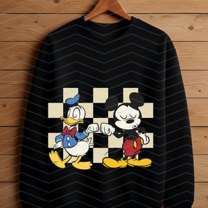 May include: Black sweatshirt featuring a graphic of Donald Duck and Mickey Mouse bumping fists. The characters are set against a checkered background. Donald Duck is wearing a blue sailor suit and Mickey Mouse is wearing red shorts.
