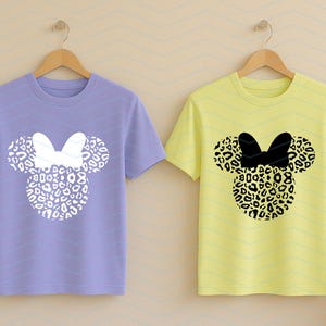 May include: Two short-sleeved t-shirts on wooden hangers. One is lavender with a white bow and leopard print mouse ears. The other is yellow with a black bow and leopard print mouse ears. The shirts are a fun, playful design.