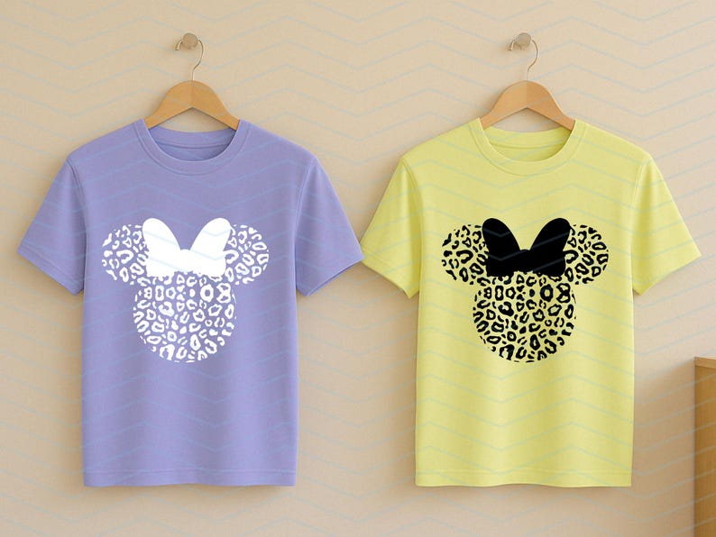 May include: Two short-sleeved t-shirts on wooden hangers. One is lavender with a white bow and leopard print mouse ears. The other is yellow with a black bow and leopard print mouse ears. The shirts are a fun, playful design.