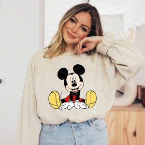 May include: A beige sweatshirt featuring a cartoon image of Mickey Mouse sitting down with his arms outstretched, wearing red shorts and yellow shoes.