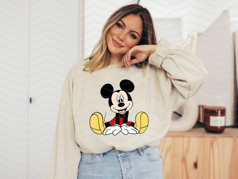 May include: A beige sweatshirt featuring a cartoon image of Mickey Mouse sitting down with his arms outstretched, wearing red shorts and yellow shoes.