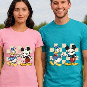 May include: Pink and teal t-shirts featuring a cartoon design of Mickey Mouse and Donald Duck. The characters are shown fist-bumping on a checkered background. The shirts are short-sleeved and worn by two people.