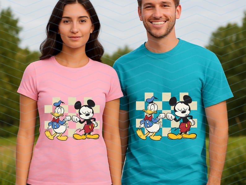 May include: Pink and teal t-shirts featuring a cartoon design of Mickey Mouse and Donald Duck. The characters are shown fist-bumping on a checkered background. The shirts are short-sleeved and worn by two people.