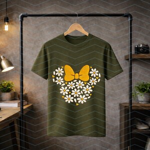 Puede incluir: Camiseta verde oliva con un dise&ntilde;o floral en forma de lazo y orejas de rat&oacute;n. El lazo es amarillo y las flores son blancas con centros amarillos. La camiseta est&aacute; colgada en una percha negra.