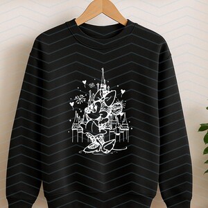 May include: Black crewneck sweatshirt featuring a white line drawing of Minnie Mouse in front of a castle. The design includes fireworks and hearts. The sweatshirt is hanging on a wooden hanger.