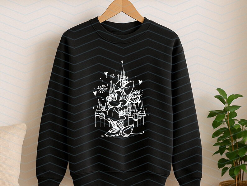 May include: Black crewneck sweatshirt featuring a white line drawing of Minnie Mouse in front of a castle. The design includes fireworks and hearts. The sweatshirt is hanging on a wooden hanger.
