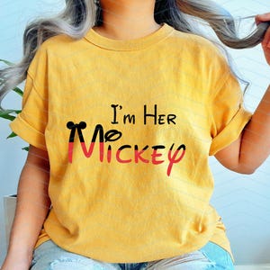 Op de afbeelding: Geel T-shirt met zwarte en rode Mickey Mouse oren en de tekst "I'm Her Mickey" gedrukt op de voorkant.