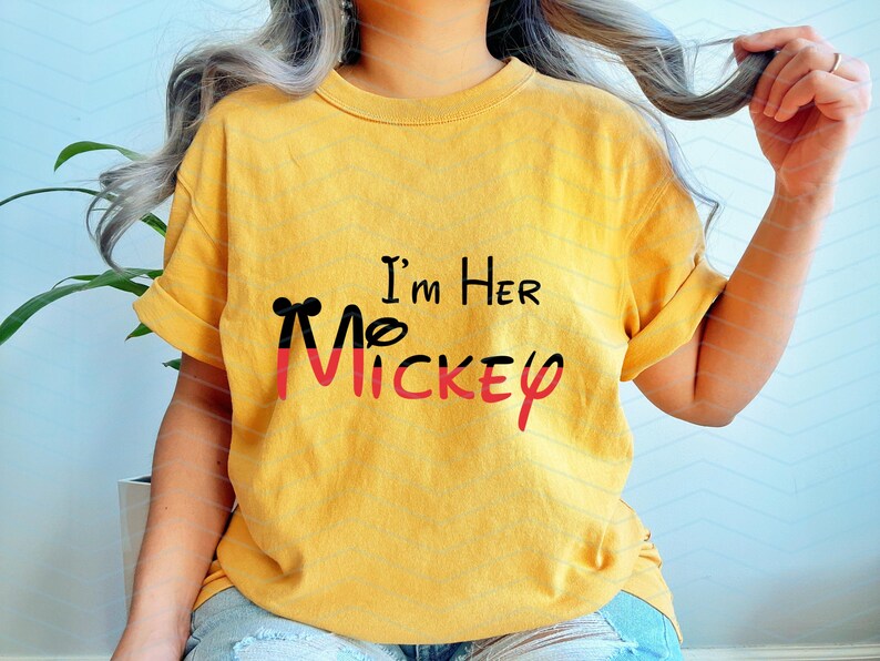 Op de afbeelding: Geel T-shirt met zwarte en rode Mickey Mouse oren en de tekst "I'm Her Mickey" gedrukt op de voorkant.