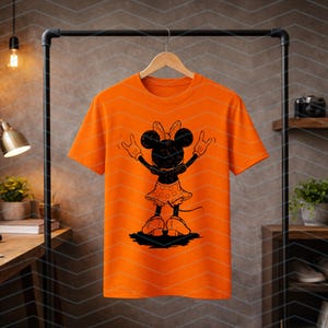 May include: Bright orange t-shirt featuring a black silhouette of Minnie Mouse with her arms raised. Minnie Mouse is wearing a polka-dot skirt and bow. The t-shirt is hanging on a black clothing rack.