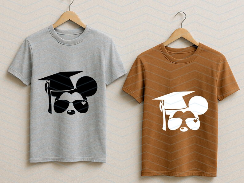 May include: Two short-sleeved t-shirts on wooden hangers. One is gray, the other is brown. Each shirt features a graphic of a mouse head wearing a graduation cap and sunglasses with heart-shaped lenses.
