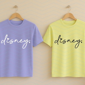 Op de afbeelding: Twee shirts met korte mouwen, &eacute;&eacute;n lavendelkleurig en &eacute;&eacute;n geel, elk met het woord "disney" in een cursief lettertype. Het gele shirt heeft een klein hartje boven de "i". Beide shirts hangen aan houten hangers.