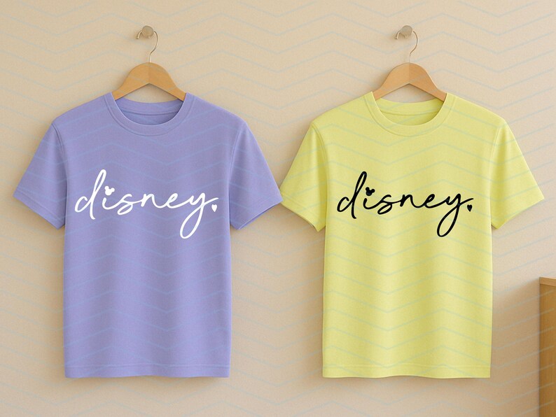 Op de afbeelding: Twee shirts met korte mouwen, &eacute;&eacute;n lavendelkleurig en &eacute;&eacute;n geel, elk met het woord "disney" in een cursief lettertype. Het gele shirt heeft een klein hartje boven de "i". Beide shirts hangen aan houten hangers.