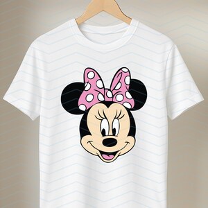May include: White t-shirt featuring a cartoon graphic of Minnie Mouse's face. The design includes black ears, a pink bow with white polka dots, and a smiling face. The shirt is hanging on a wooden hanger.