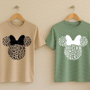 May include: Two short-sleeved t-shirts hanging on wooden hangers. One is tan with a black bow and leopard print mouse head design. The other is green with a white bow and leopard print mouse head design. The shirts are displayed against a light beige background.