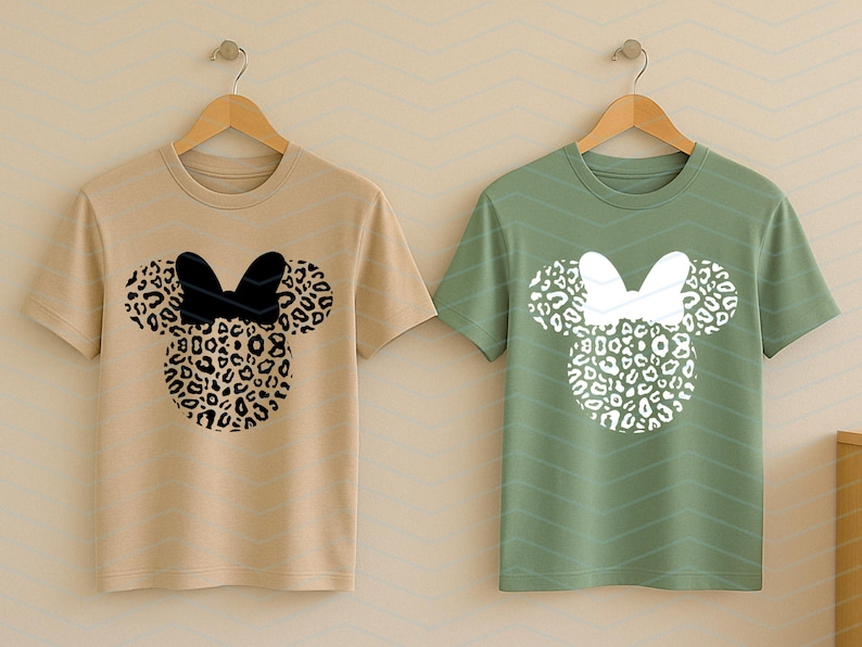 May include: Two short-sleeved t-shirts hanging on wooden hangers. One is tan with a black bow and leopard print mouse head design. The other is green with a white bow and leopard print mouse head design. The shirts are displayed against a light beige background.