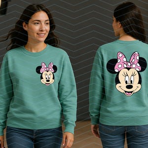 May include: Teal sweatshirt featuring Minnie Mouse. The front displays Minnie's face with a pink polka dot bow. The back of the sweatshirt shows Minnie's face and bow. The sweatshirt is a crew neck style and is paired with blue jeans.