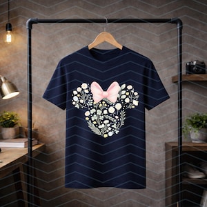 May include: Navy blue t-shirt featuring a floral design shaped like a mouse head, with a pink bow. The design includes white and yellow flowers and green leaves. The t-shirt is hanging on a wooden hanger.