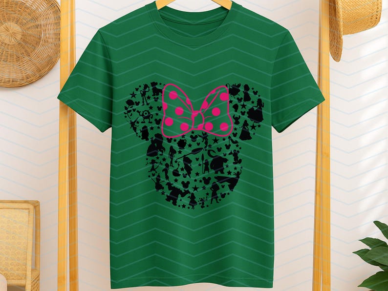 May include: A dark green short-sleeve t-shirt featuring a subtle horizontal chevron pattern. The center displays a large black silhouette of a mouse head shape filled with various small character figures and stars, topped with a bright pink polka-dot bow.