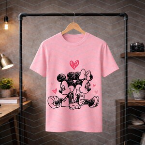 Peut inclure: T-shirt rose clair avec une illustration en noir et blanc de Mickey et Minnie Mouse enlac&eacute;s, surmont&eacute;s d'un c&oelig;ur. Le motif est centr&eacute;, avec de petits c&oelig;urs dispers&eacute;s autour des personnages.