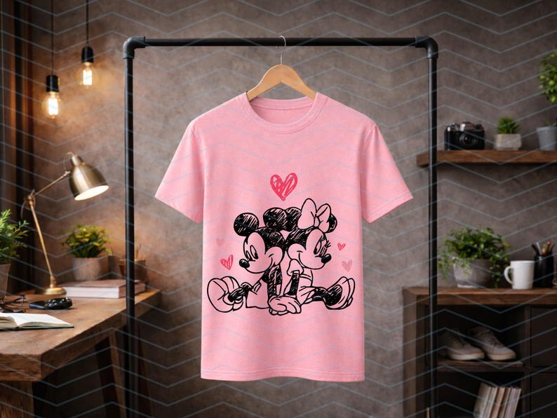 Peut inclure: T-shirt rose clair avec une illustration en noir et blanc de Mickey et Minnie Mouse enlac&eacute;s, surmont&eacute;s d'un c&oelig;ur. Le motif est centr&eacute;, avec de petits c&oelig;urs dispers&eacute;s autour des personnages.