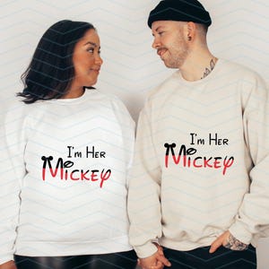 Op de afbeelding: Een stel draagt witte sweatshirts met zwarte tekst die "I'm Her Mickey" en "I'm Her Mickey" zegt met een rode Mickey Mouse-kop grafiek. De vrouw draagt een wit sweatshirt en de man draagt een cr&egrave;mekleurig sweatshirt.