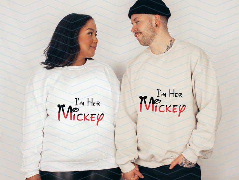 Op de afbeelding: Een stel draagt witte sweatshirts met zwarte tekst die "I'm Her Mickey" en "I'm Her Mickey" zegt met een rode Mickey Mouse-kop grafiek. De vrouw draagt een wit sweatshirt en de man draagt een cr&egrave;mekleurig sweatshirt.
