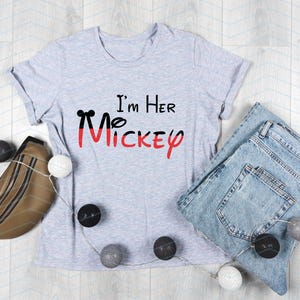 Op de afbeelding: Een grijs T-shirt met de tekst "I'm Her Mickey" in zwarte en rode letters. Het shirt ligt plat op een houten oppervlak met een bruine leren heuptas, blauwe jeans en witte en grijze decoratieve ballen.