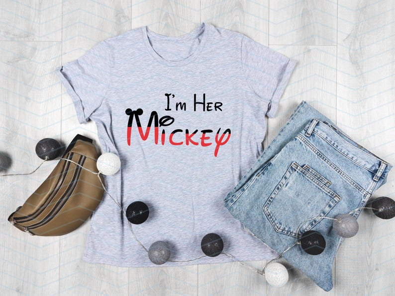 Op de afbeelding: Een grijs T-shirt met de tekst "I'm Her Mickey" in zwarte en rode letters. Het shirt ligt plat op een houten oppervlak met een bruine leren heuptas, blauwe jeans en witte en grijze decoratieve ballen.