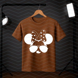 May include: A brown t-shirt featuring a white graphic of a cartoon mouse face with sunglasses, a polka dot bow, and large round ears. The t-shirt is hanging on a wooden hanger against a dark background.