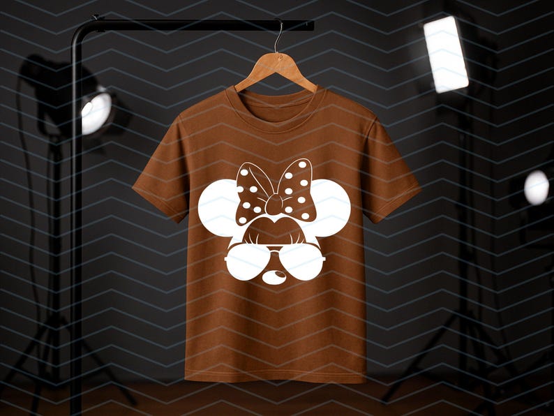 May include: A brown t-shirt featuring a white graphic of a cartoon mouse face with sunglasses, a polka dot bow, and large round ears. The t-shirt is hanging on a wooden hanger against a dark background.