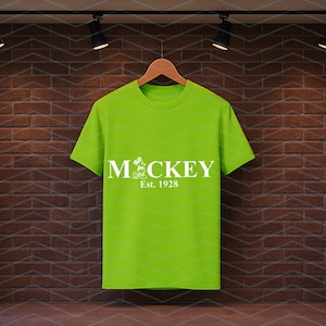 Pu&ograve; includere: Una maglietta verde lime con la scritta "MCKEY" in bianco, con un personaggio dei cartoni animati e la scritta "Est. 1928". La maglietta &egrave; appesa a una gruccia di legno su uno sfondo di muro di mattoni.