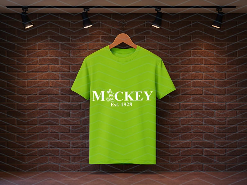 Pu&ograve; includere: Una maglietta verde lime con la scritta "MCKEY" in bianco, con un personaggio dei cartoni animati e la scritta "Est. 1928". La maglietta &egrave; appesa a una gruccia di legno su uno sfondo di muro di mattoni.