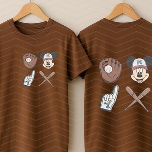 May include: Two brown t-shirts with a baseball theme featuring Mickey Mouse. One shirt has Mickey Mouse holding a baseball glove and the other has Mickey Mouse holding a baseball bat and a number one finger.