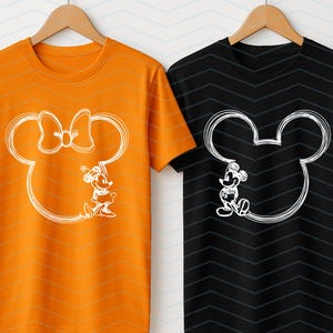 Puede incluir: Dos camisetas colgadas en perchas de madera. Una es naranja con un contorno blanco de Minnie Mouse y la otra es negra con un contorno blanco de Mickey Mouse. Ambas camisetas tienen un dise&ntilde;o de dibujos animados.