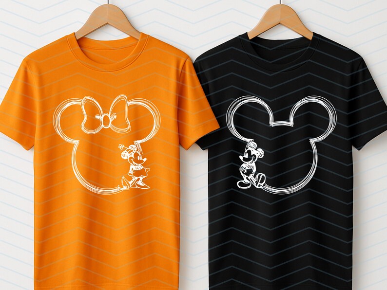 Puede incluir: Dos camisetas colgadas en perchas de madera. Una es naranja con un contorno blanco de Minnie Mouse y la otra es negra con un contorno blanco de Mickey Mouse. Ambas camisetas tienen un dise&ntilde;o de dibujos animados.