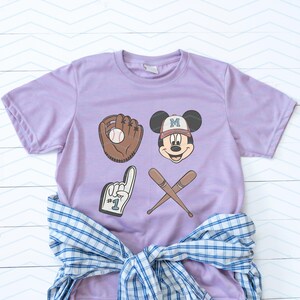 May include: A lavender t-shirt with a baseball glove, baseball, crossed bats, and a Mickey Mouse character wearing a baseball cap with the letter "M" on it. The Mickey Mouse character is wearing a light blue baseball cap with a white "M" on it. The t-shirt has the text "#1" on the glove.
