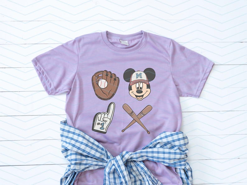 May include: A lavender t-shirt with a baseball glove, baseball, crossed bats, and a Mickey Mouse character wearing a baseball cap with the letter "M" on it. The Mickey Mouse character is wearing a light blue baseball cap with a white "M" on it. The t-shirt has the text "#1" on the glove.