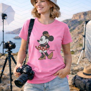 K&ouml;nnte beinhalten: Ein pinkfarbenes T-Shirt mit einer Minnie Mouse-Grafik. Die Zeichentrickfigur tr&auml;gt einen gepunkteten Rock und eine Schleife. Das Wort "Disney" ist darunter gedruckt. Die Person h&auml;lt eine Kamera.