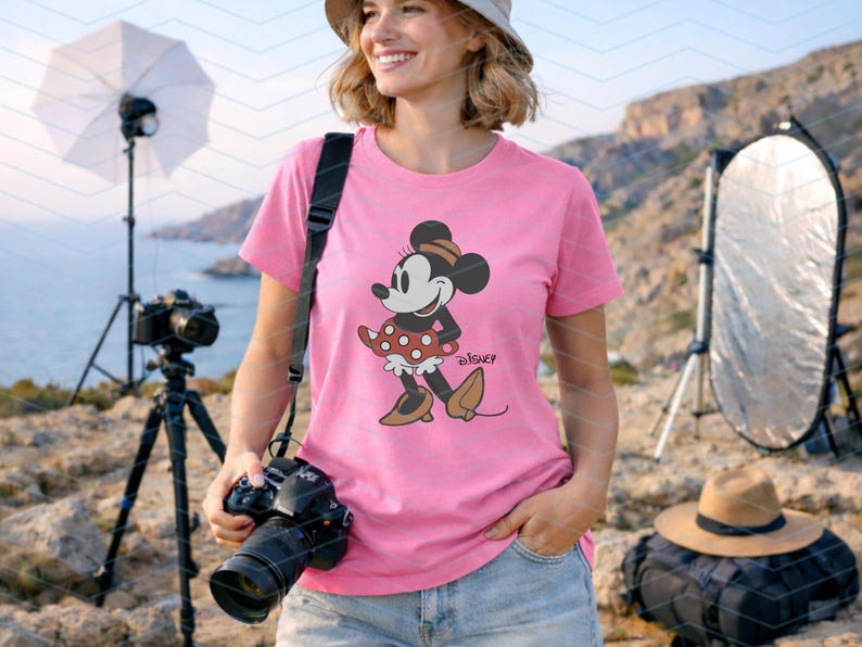 K&ouml;nnte beinhalten: Ein pinkfarbenes T-Shirt mit einer Minnie Mouse-Grafik. Die Zeichentrickfigur tr&auml;gt einen gepunkteten Rock und eine Schleife. Das Wort "Disney" ist darunter gedruckt. Die Person h&auml;lt eine Kamera.